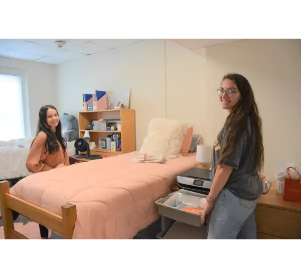 Two students stand and smile inside a residence hall room in SHSU’s Elliot Hall. The room features a neatly made bed with a peach comforter, a desk with organized supplies and decorations, and a window letting in natural light.