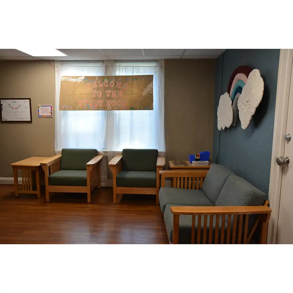 One of the study rooms in SHSU’s Elliott Hall features wooden furniture with green cushions, including chairs and a small couch arranged around wooden side tables. A banner reading “Welcome to the Study Nook” hangs above two windows, and a wall decoration shaped like a rainbow with fluffy clouds adds a cozy touch to the room.