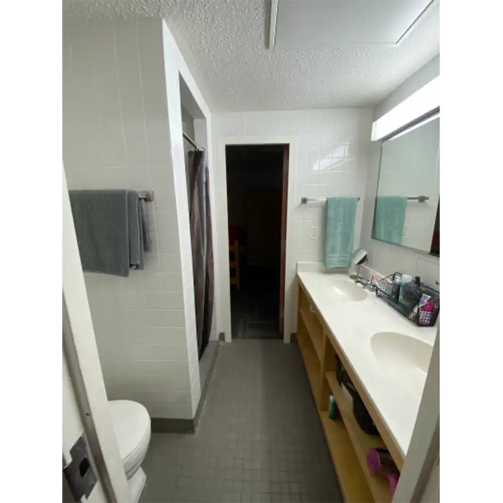 Bathroom in SHSU’s Estill Hall with dual sinks, mirror, toilet, and shower area. Countertop holds toiletries; shelves below store towels.