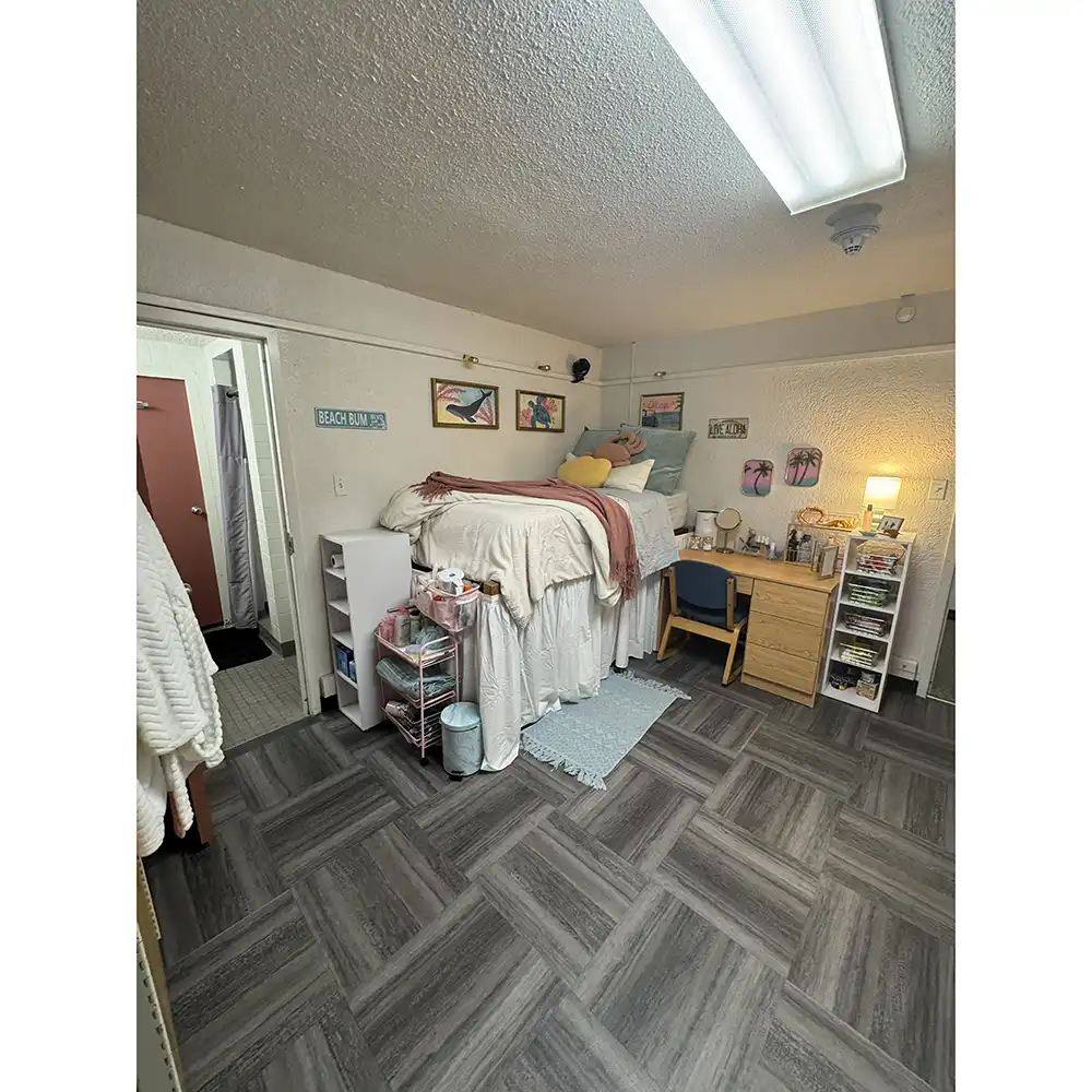 A decorated bedroom in SHSU’s Estill Hall featuring a raised bed with white and maroon bedding, a cozy rug, and a desk with a lamp and organized supplies. The room includes wall art, storage bins, and an open doorway leading to a bathroom. The space has dark patterned flooring and soft lighting, giving it a comfortable, personalized feel.