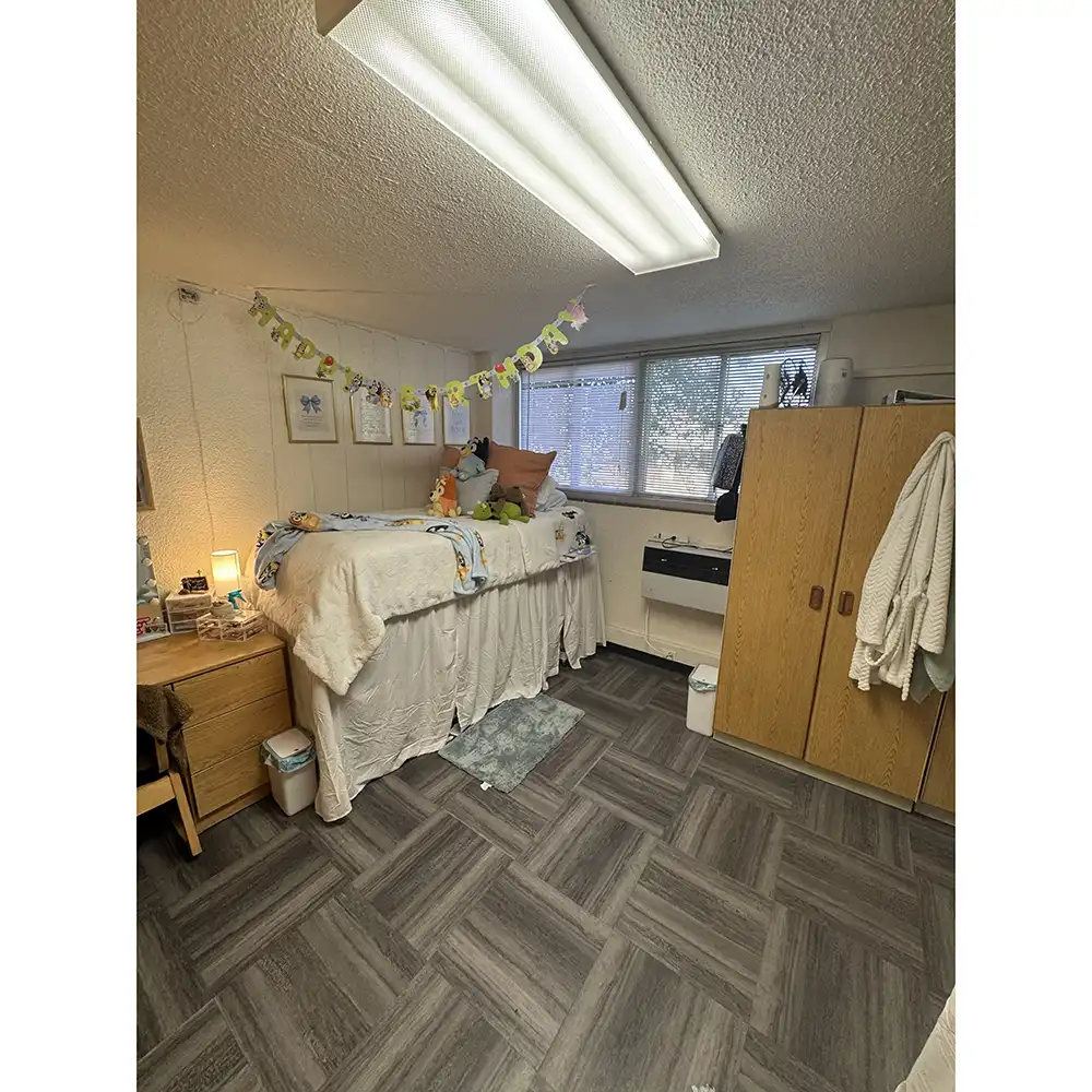 Bedroom in SHSU’s Estill Hall with an elevated single bed, nightstand, wardrobe, and window with blinds. Room includes personal decor and tiled flooring.