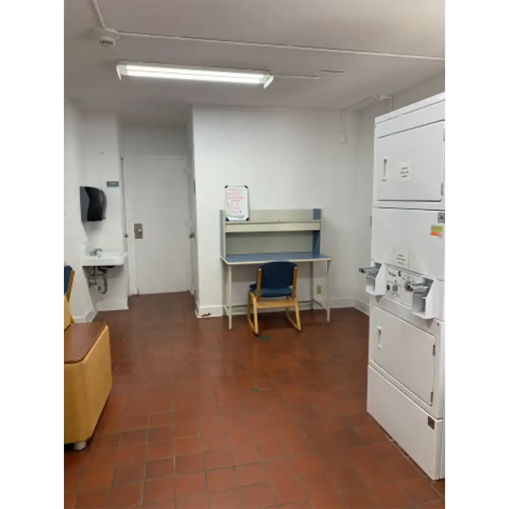 Laundry room in SHSU’s Estill Hall with stacked white washers and dryers, tiled floor, white walls, a desk with a blue chair, and a wall-mounted sink in the corner. Fluorescent lights illuminate the space.