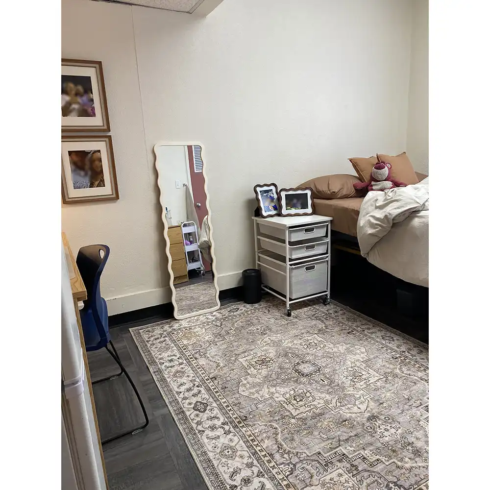 Bedroom in SHSU’s Jackson-Shaver Hall with a bed featuring beige bedding, pillows, and a stuffed animal. A white three-drawer storage unit beside the bed holds framed photos and a black cylindrical item. A wavy full-length mirror leans against the wall, and a desk with a blue chair sits nearby. The room has dark wood-style flooring and a beige and gray patterned area rug.