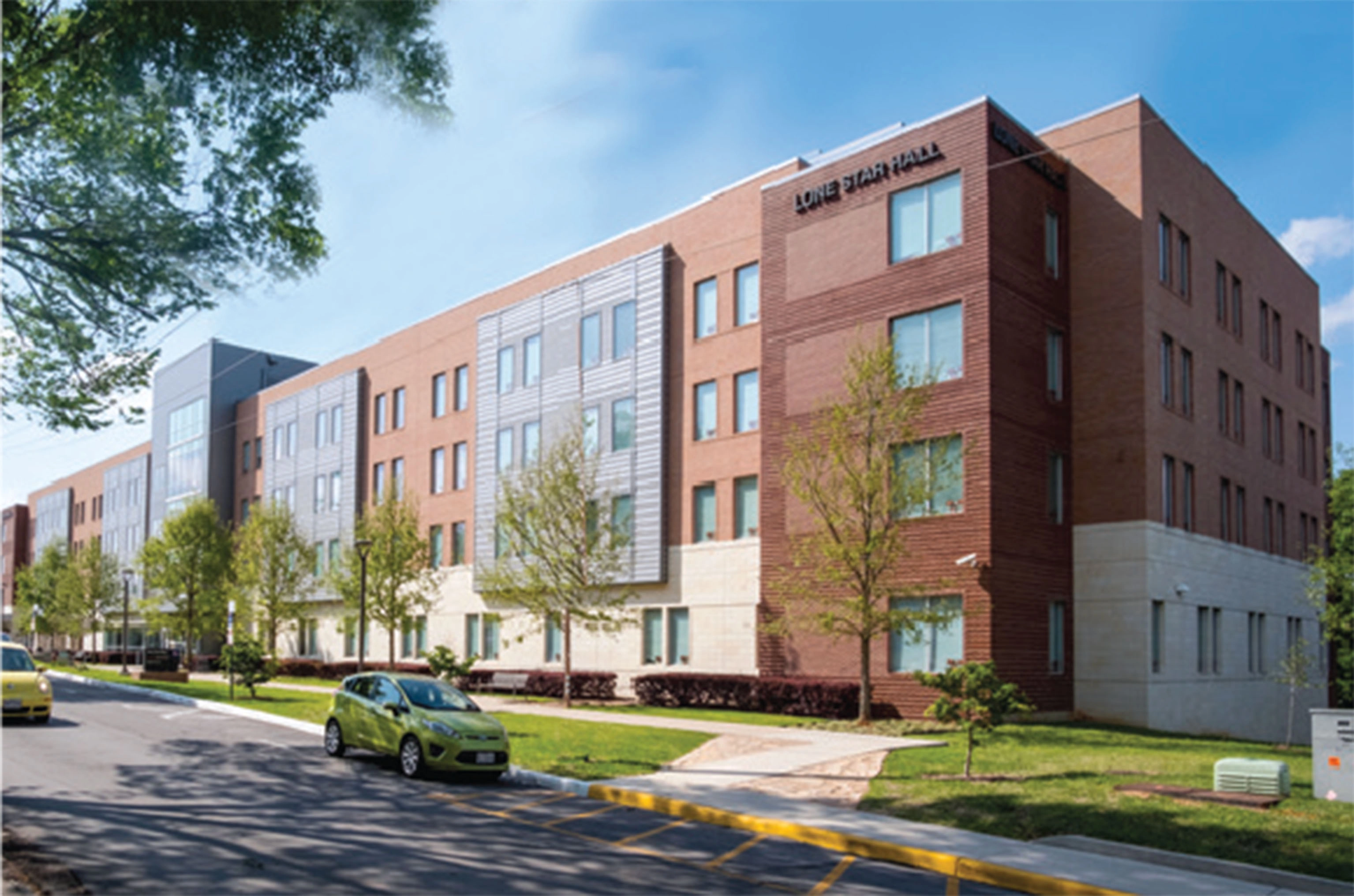 Modern four-story residence hall named "Lone Star Hall," featuring a mix of red brick, light stone, and silver metal paneling. The building is lined with large windows and surrounded by young trees and a landscaped lawn, with cars parked along the street in front.