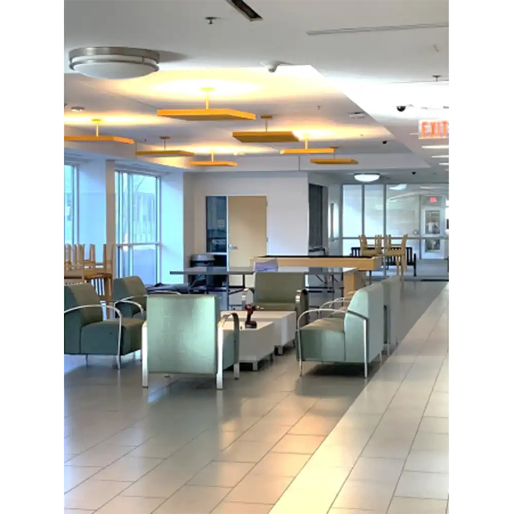 Spacious lobby in SHSU’s Lone Star Hall with green cushioned chairs and white square tables, yellow rectangular ceiling lights, large windows providing natural light, and additional seating areas in the background. The floor is light-colored tile, contributing to the bright, modern atmosphere.
