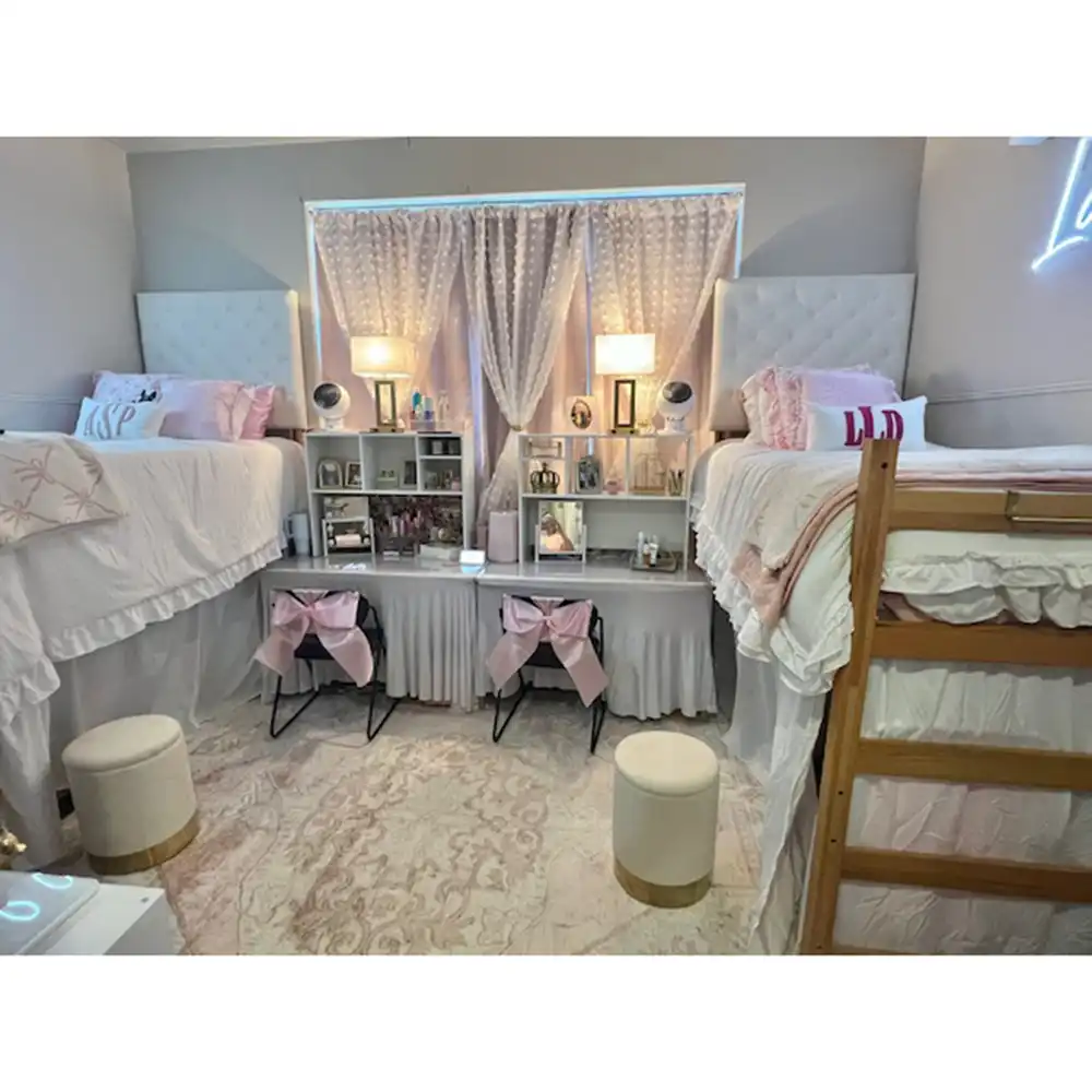 A decorated Piney Woods Hall bedroom at SHSU featuring two lofted beds with white ruffled bedding and pink accent pillows. Between the beds are shelves holding lamps, framed photos, and small mirrors. Beneath the shelves are two desks with black chairs tied with pink bows. Two round white stools with gold bases and a patterned rug complete the cozy, coordinated setup.