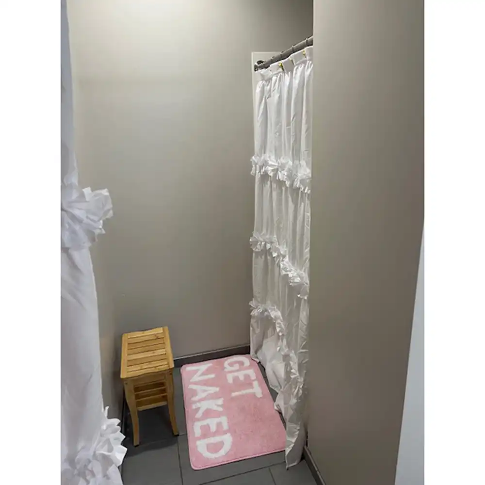 Shower area in SHSU's Piney Woods Hall featuring a white ruffled shower curtain on a rod, partially open to reveal the shower interior. A pink bath mat with the words 'GET NAKED' in white letters is placed in front of the shower. A small wooden stool sits to the left of the mat. The walls are light gray, and the floor is tiled in dark gray.