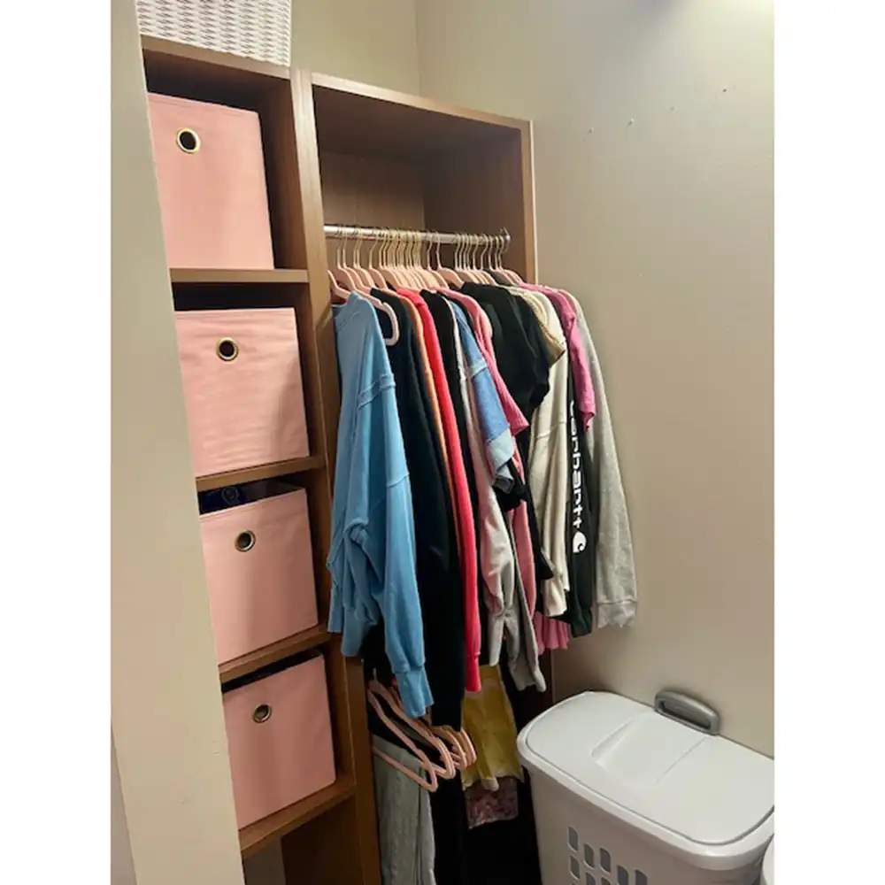 Storage area in SHSU's Piney Woods Hall featuring a wooden shelving unit with three pink fabric storage bins on the left. A clothing rack holds various shirts and hoodies on wooden hangers in colors like blue, red, gray, and black. Additional empty hangers are below the clothes. A white plastic laundry basket with a lid is on the right. The closet walls are beige.