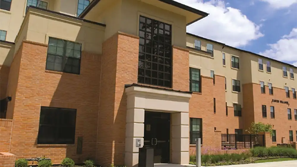 Exterior view of Raven Village at Sam Houston State University. The building has beige and brick walls with multiple rectangular windows. The central entrance features a glass panel above the doorway. The words 'Raven Village' are displayed on the right side of the building. The sky is partly cloudy, and landscaped greenery surrounds the entrance.