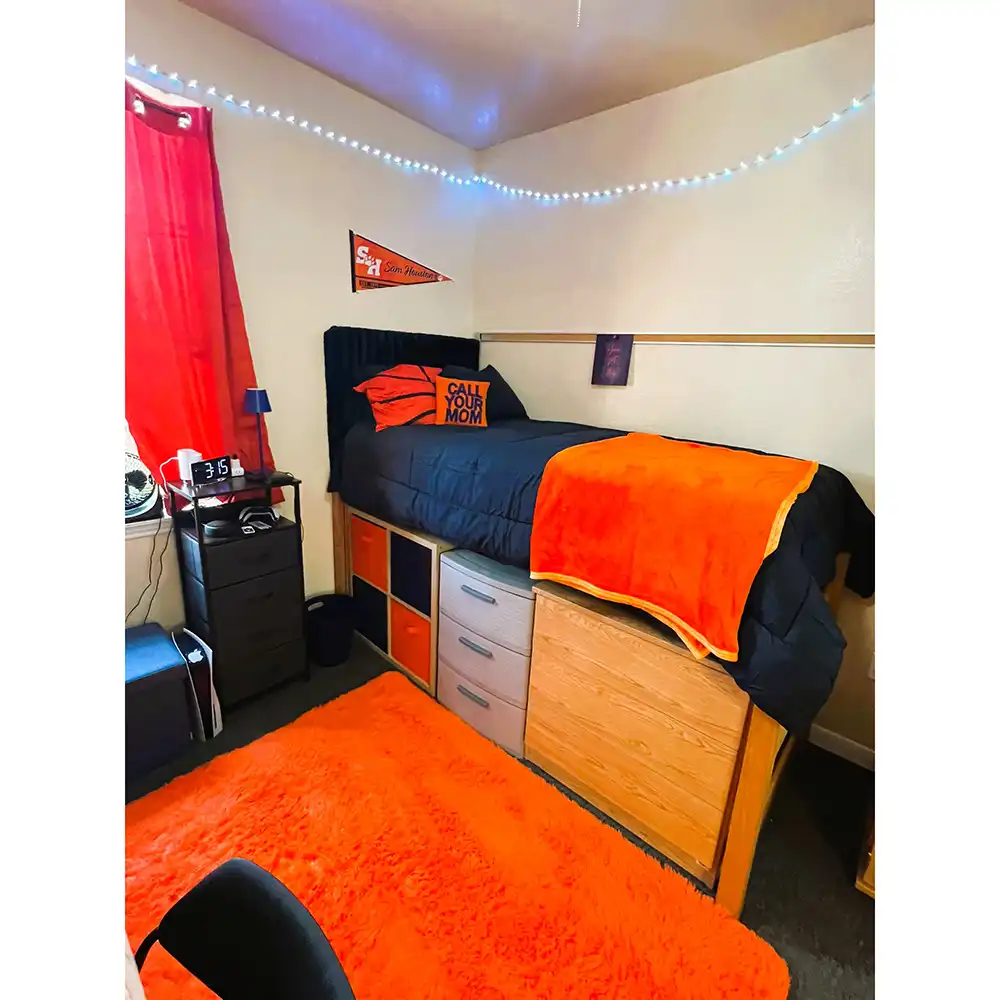 Bedroom in SHSU's Raven Village featuring a lofted bed with navy blue and orange bedding, storage drawers and cubbies underneath, battery operated string lights along the ceiling, an orange curtain on the window, and an orange rug on the floor. A small nightstand holds a lamp, alarm clock, and personal items, with an orange pennant flag mounted on the wall above the bed.
