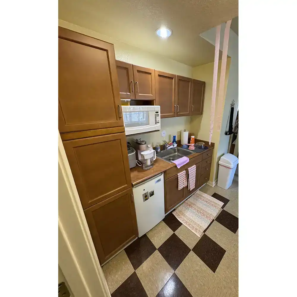 Kitchenette in SHSU's Raven Village features wooden cabinets with silver handles, a microwave mounted under upper cabinets, and a small refrigerator integrated into the lower cabinets. The countertop holds a coffee maker, small ice machine, and a dish drying rack beside the sink. Dish towels hang from the oven handle, and two rugs cover the checkered tile floor. A trash can is placed near the entrance to an adjacent room or hallway.
