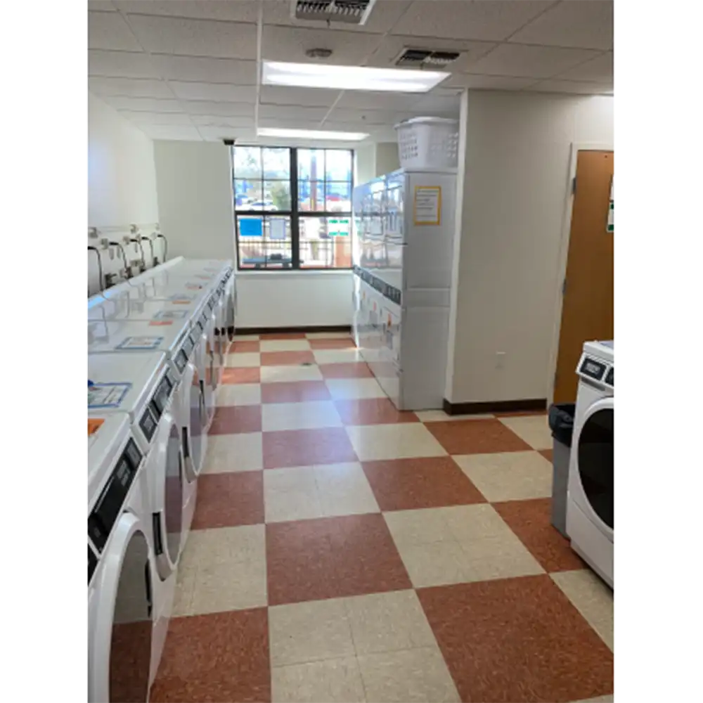 Laundry room in SHSU's Raven Village featuring a checkered floor with red and beige tiles, rows of front-loading washers and dryers on both sides, a window at the far end providing natural light and an outdoor view, recessed ceiling lights, and a laundry basket placed on one of the machines.