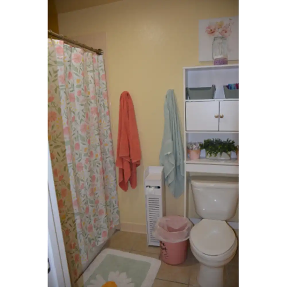 Bathroom in SHSU's Raven Village featuring a shower with a floral-patterned curtain, an orange towel and a light blue towel hanging on the wall, a white shelving unit above the toilet holding a plant and toiletries, a pink trash bin beside the toilet, and a decorative rug on the floor. The walls are painted light yellow.