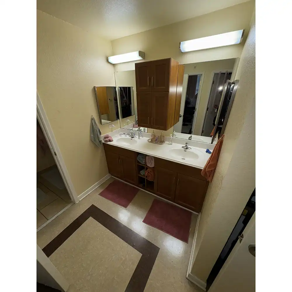 Sink/vanity area in SHSU's Raven Village featuring a double sink with a white countertop and wooden cabinets below. Two mirrors with fluorescent lights are mounted above the sinks, with a wooden storage cabinet between them. Red bath mats are placed in front of each sink, and towels hang on both sides—gray on the left and orange on the right. The room has beige walls and a tiled floor with a brown square pattern near the entrance to an adjacent room.