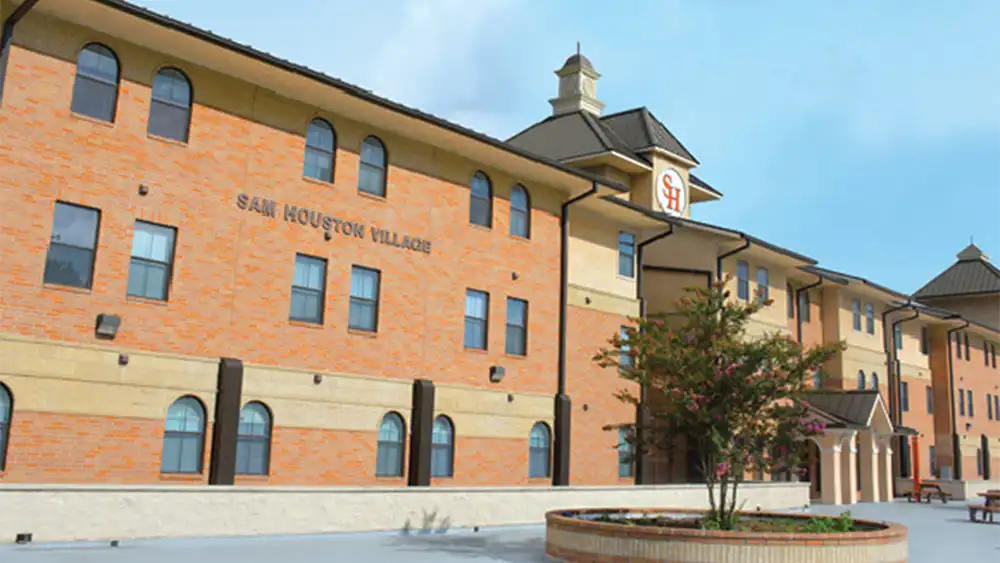 Image Alt Text   Exterior view of Sam Houston Village at Sam Houston State University. The three-story residential building features red brick walls with beige stone accents and multiple windows, including arched ones on the ground floor. 'SAM HOUSTON VILLAGE' is displayed in large letters on the side of the building. A small courtyard in front includes a circular brick planter with a tree and surrounding greenery. The sky is mostly clear with a few clouds.