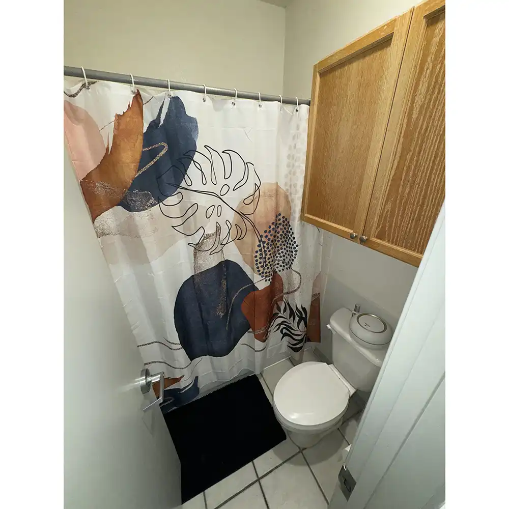 Bathroom at SHSU's Sam Houston Village featuring a white shower curtain with abstract leaf and plant designs in blue, orange, and beige. A white toilet with a closed lid sits next to a wall-mounted wooden cabinet. The floor is white-tiled with a black bath mat in front of the shower.