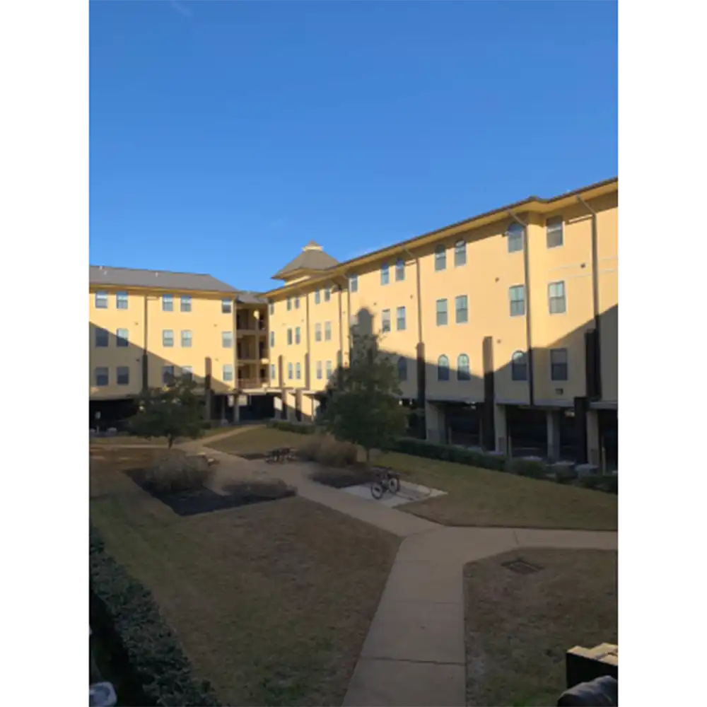 Courtyard at Sam Houston Village, SHSU, surrounded by a three-story yellow-beige building with many windows. The grassy area features pathways, small trees, shrubs, benches, and a bicycle. The sky is clear and blue, suggesting a sunny day.