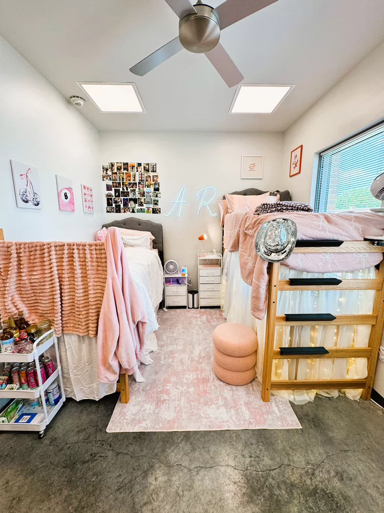 Shared student room in SHSU’s San Jacinto Hall featuring two XL bunkable twin beds, one lofted with storage and lights underneath, and the other at standard height. The room is decorated with pink bedding, wall art, photos, and a neon sign, with natural light coming through a window.