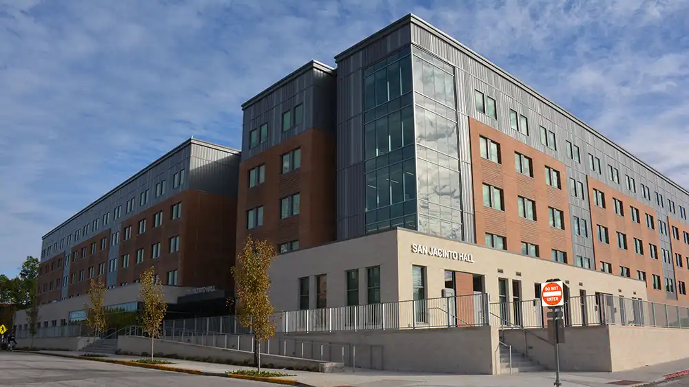 Exterior view of San Jacinto Hall at Sam Houston State University. The modern multi-story building features a brick and glass facade with large windows. It is located on a street corner where young trees line the sidewalk in front of the building. The sky is partly cloudy.