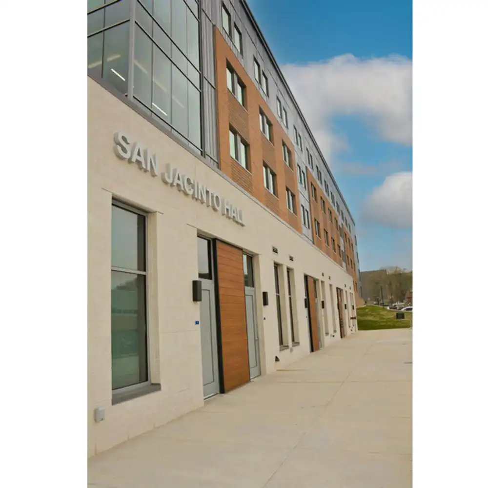 Exterior view of SHSU's San Jacinto Studio Apartments, a multi-story building with modern architecture featuring large windows and a mix of light-colored stone and brick. All studios are located on the first floor. The entrance displays a sign reading 'SAN JACINTO HALL' above the doors. A sidewalk leads to the entrance under a partly cloudy sky.