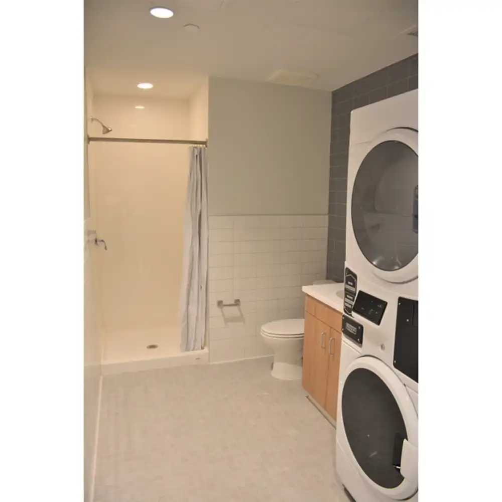 Bathroom in SHSU's San Jacinto Studio Apartments featuring a walk-in shower with a curtain, a toilet, and a wooden vanity with a white countertop. A stacked washer and dryer unit is positioned next to the vanity. The walls are partially tiled in white, and the floor is covered in light-colored tiles. Recessed ceiling lights provide illumination.