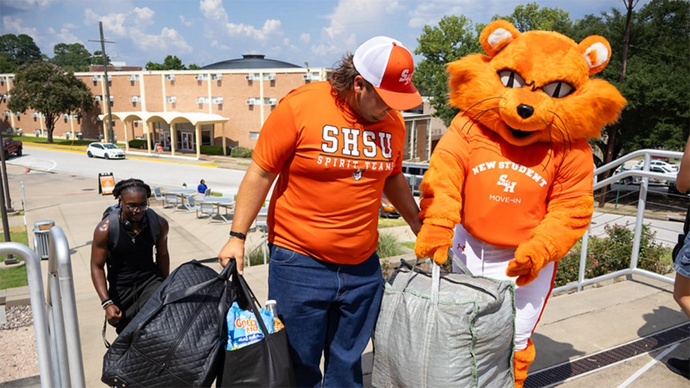 A person wearing an orange SHSU Spirit Team shirt and cap carries bags and bedding up outdoor stairs alongside Sammy Bearkat the university mascot wearing a shirt that reads "New Student Move-In." A student follows behind carrying a large black bag. Campus buildings, railings, and a roadway are visible in the background on a sunny day.