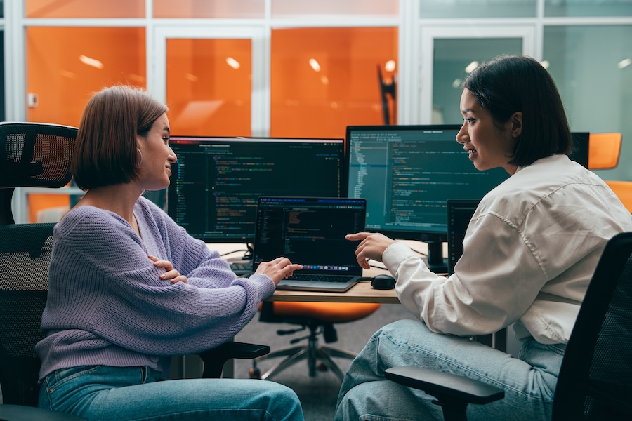 two people looking and pointing at a laptop screen with other computer screens behind