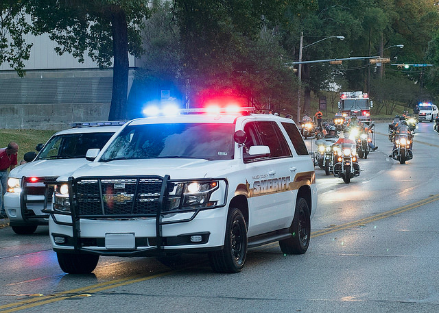 Police vans and motorcycles drive down Sam Houston Avenue during a parade.