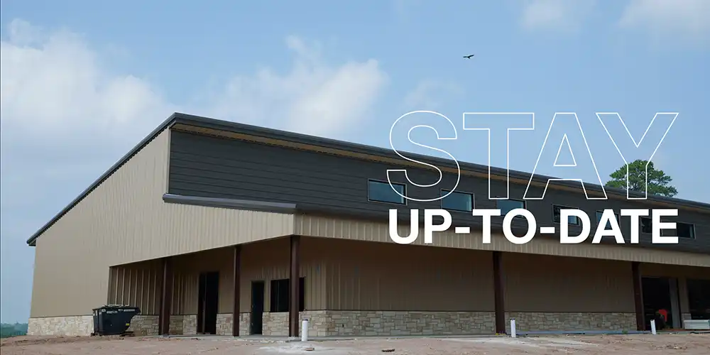 Modern warehouse-style building with beige and dark gray walls, stone brick base, and brown columns under a partly cloudy sky, with text overlay reading ‘STAY UP-TO-DATE.’