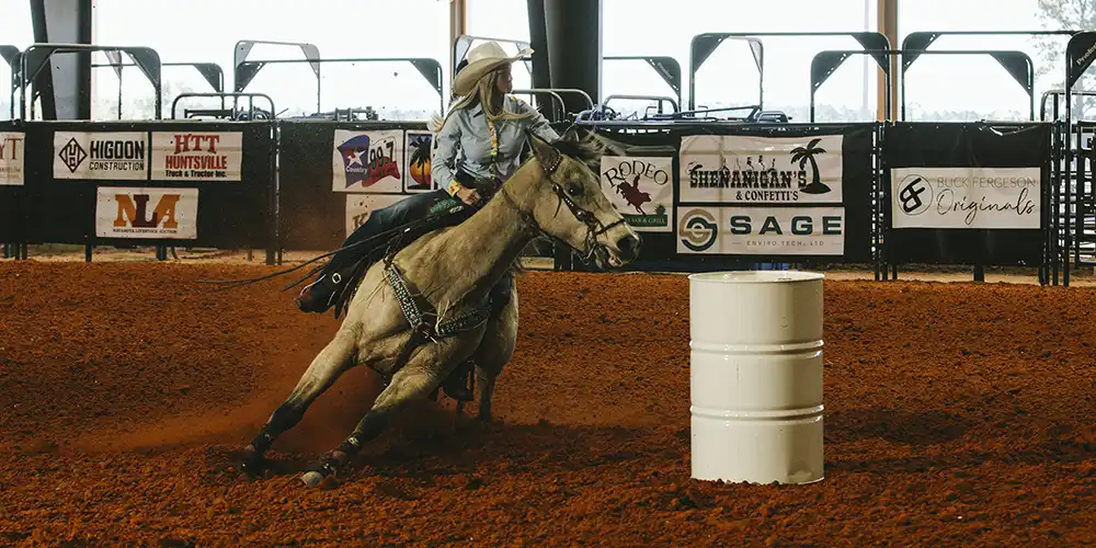 Interior of Gibbs Ranch Facility featuring a tractor driven by a person under a large metal-roofed arena with beams, and bleachers in the background.