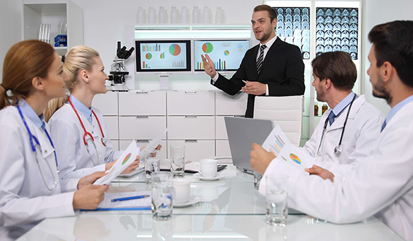4 health care professionals sitting at a table watching a man in a suit give a presentation