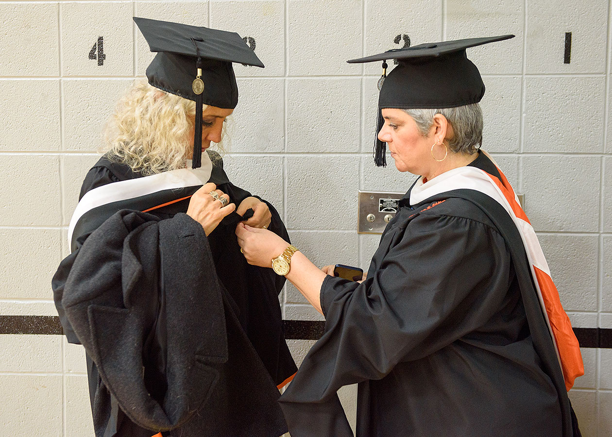 Two graduates assist each other in putting on their graduation caps and gowns.
