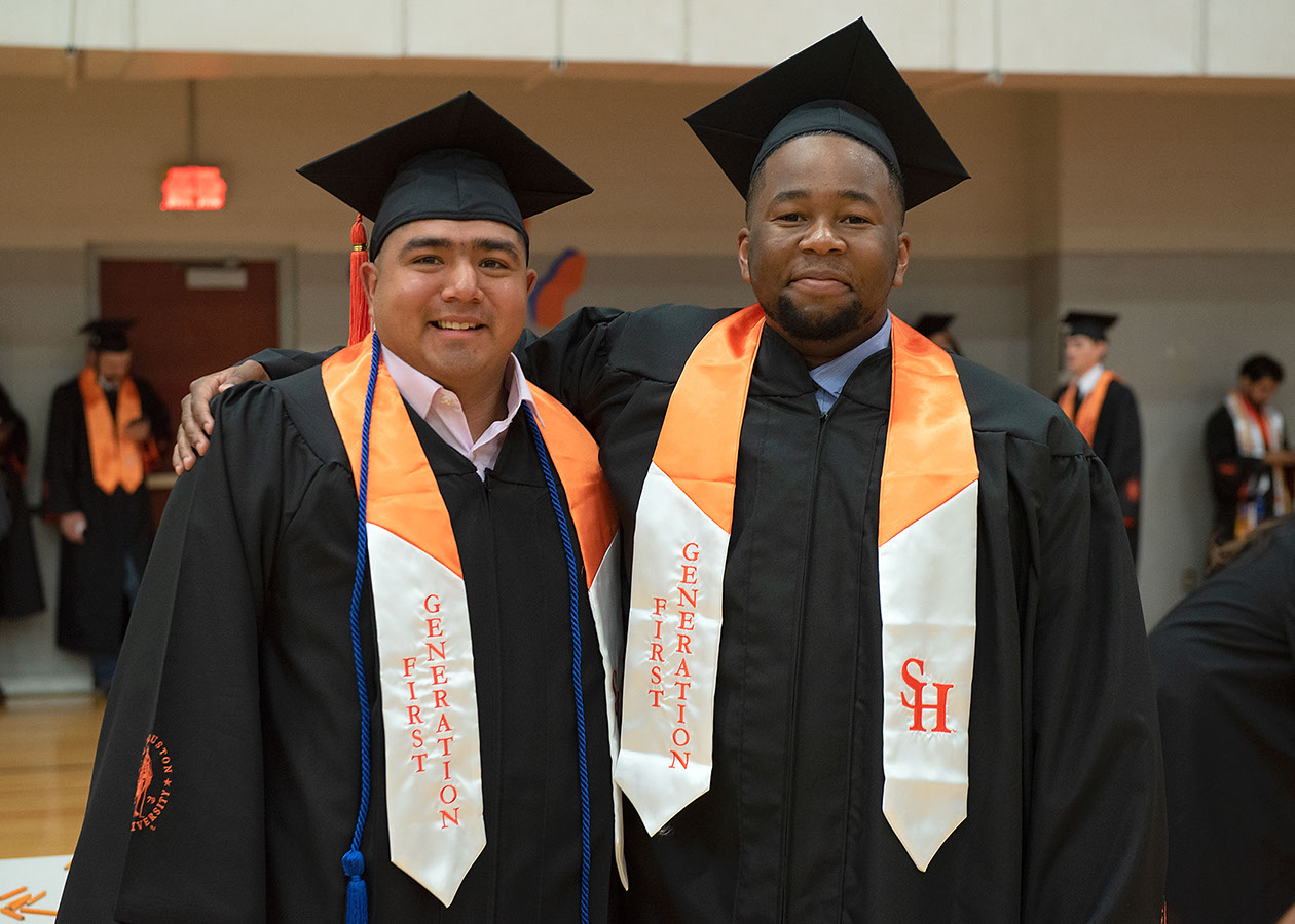 Two first-generation students stand arm-in-arm before the beginning of their commencement ceremony.