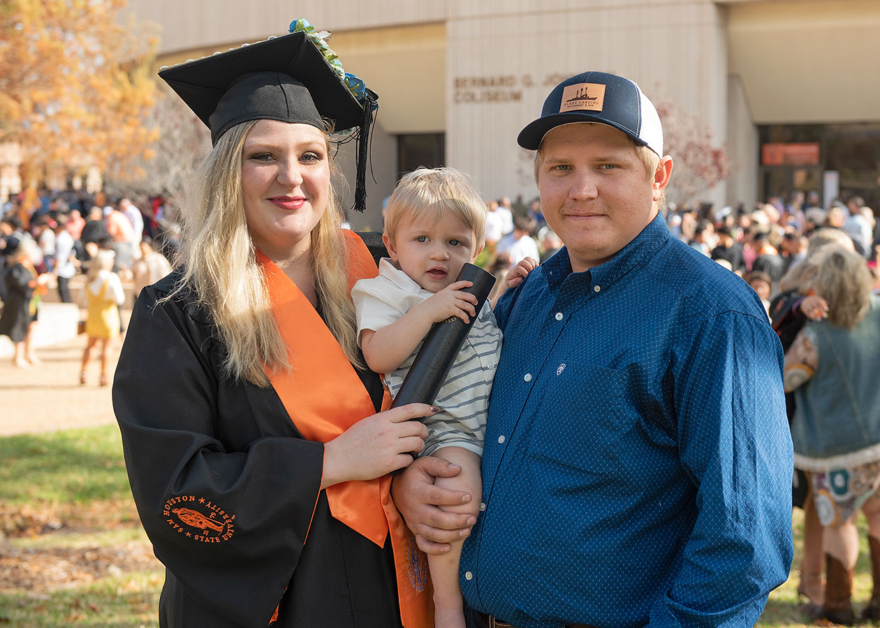 Courtney Malecek poses with her baby son and husband in her cap and gown outside of Johnson Coliseum.
