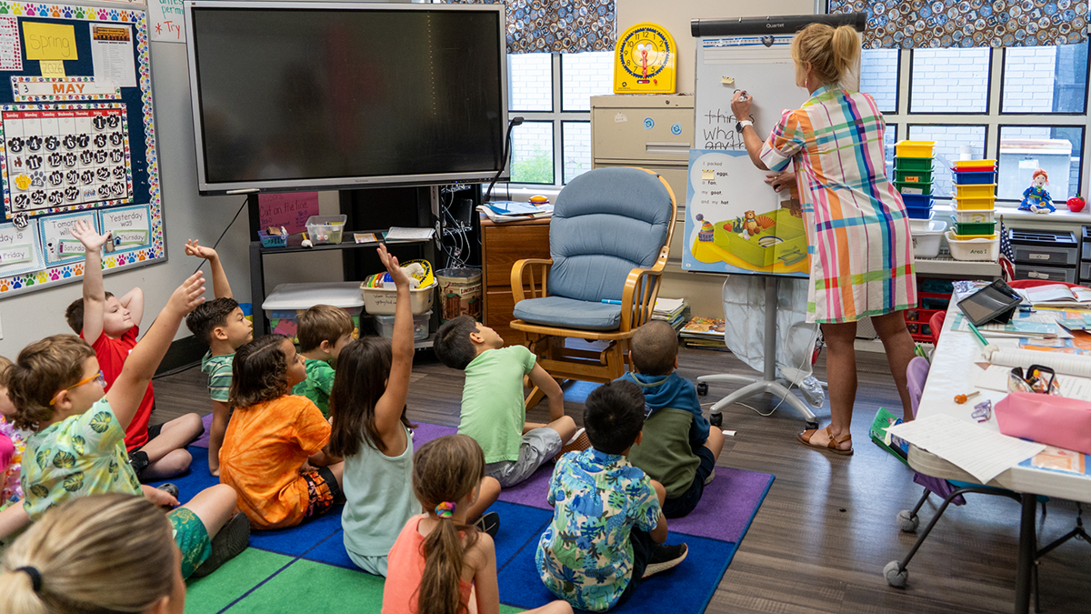 A teacher writes on a board while students look on with hands raised.