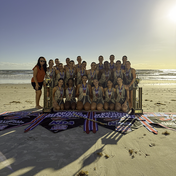 The Orange Pride Team stands on the beach with the banners and trophies that commemorate their success at this year's NDA College Nationals competition.
