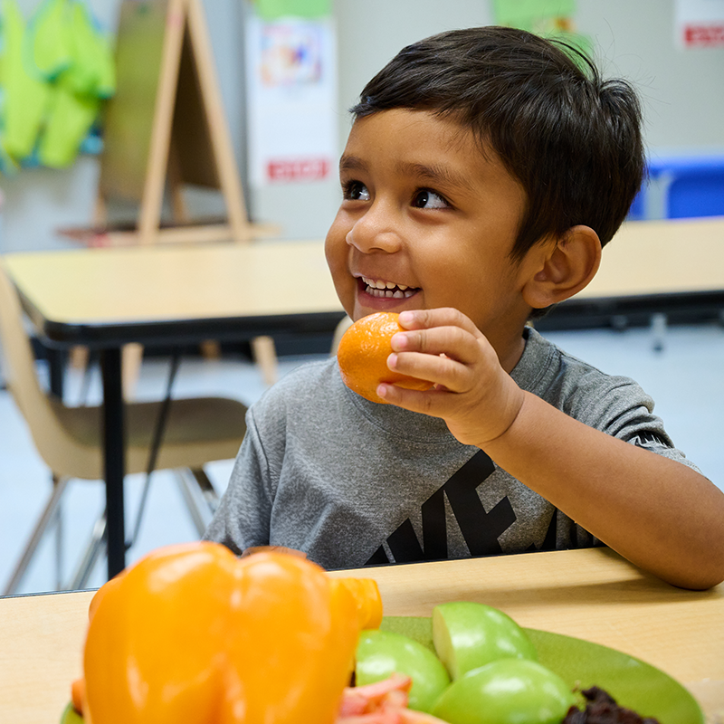 A child holds a seedless mandarin while smiling up at a dietetic intern who is just out of frame.