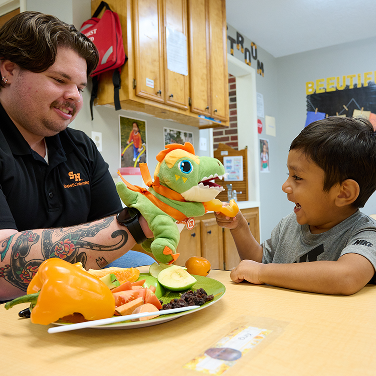 A dietetic intern uses a dinosaur puppet to play with a child they are introducing to new vegetables.