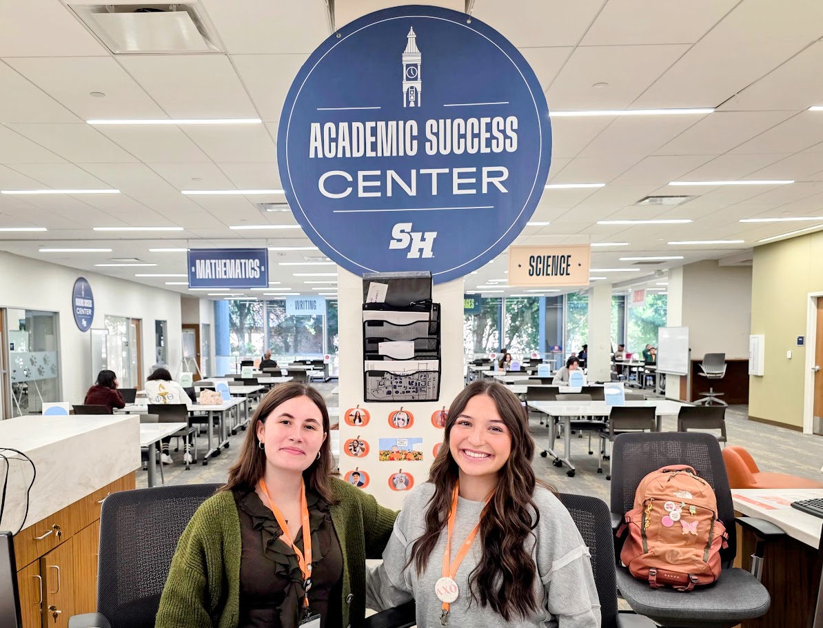 two female ambassadors sitting behind the welcome desk with the ASC sign in the rear of the pic