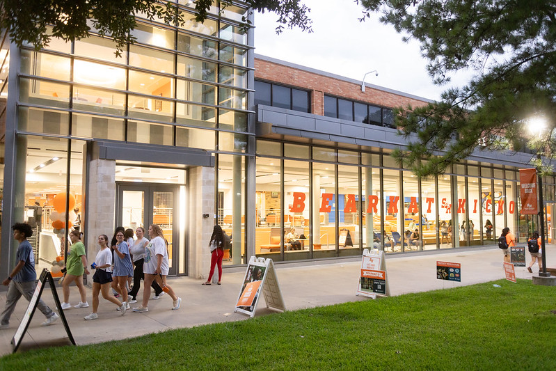people passing in front of a glass wall with orange text at dusk