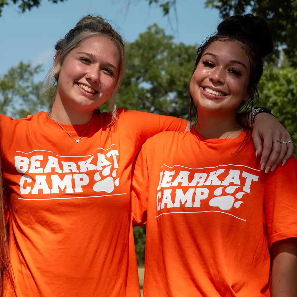Two smiling students in bright orange "Bearkat Camp" shirts stand arm-in-arm outdoors, enjoying the sun and showcasing the friendships formed during camp at Sam Houston State University.