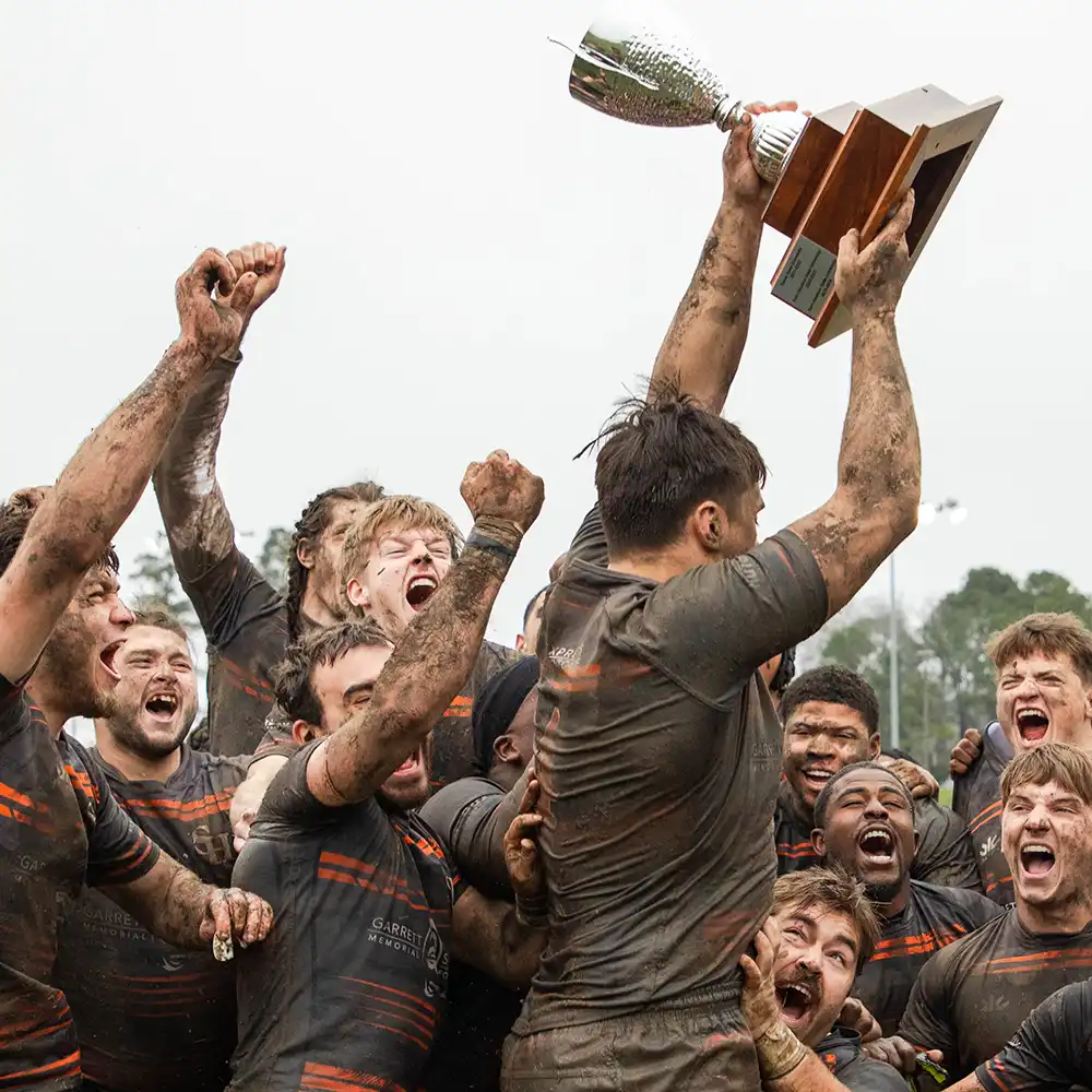Men’s rugby team grouped together celebrating their win by holding up the trophy and cheering.