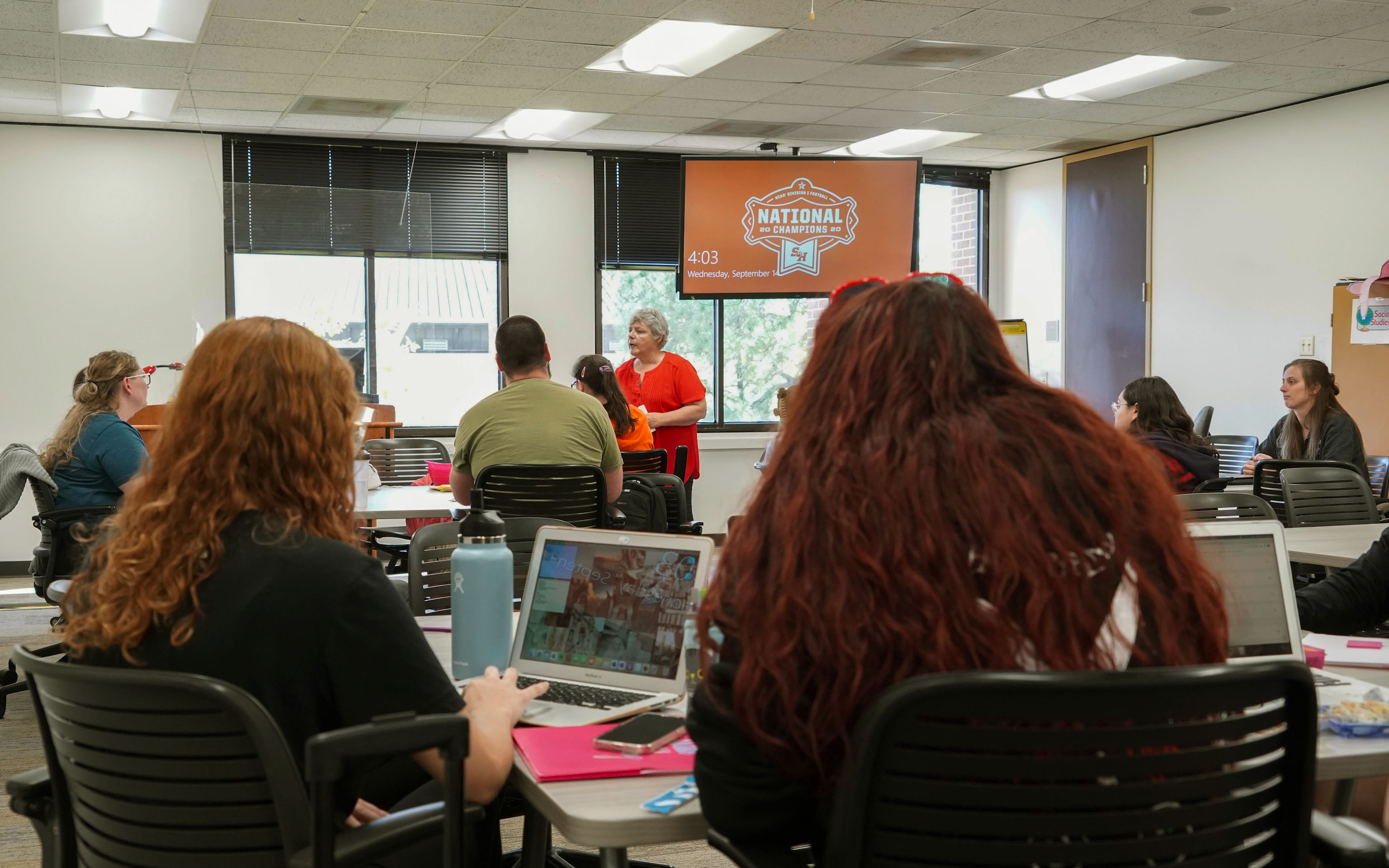 College classroom with students at desks, instructor at the front of the room giving instruction
