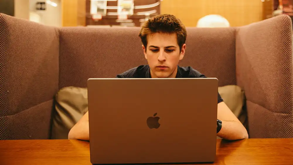 A college student sits in a booth focused on a laptop, reviewing information on the screen. The setting appears to be a quiet study area or lounge.