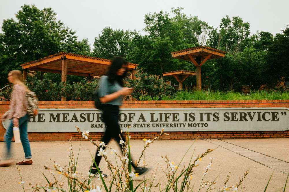 Students walk through Frank Parker Plaza on the Sam Houston State University campus, passing the wall inscribed with the university motto, “The measure of a life is its service,” surrounded by greenery and wooden pergolas.