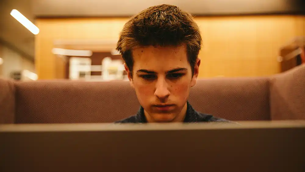 An image of a future student sitting in a cozy booth focusing intently on a laptop as he applies for housing at Sam Houston State University. The student has short brown hair, a navy-blue shirt, and a focused expression. The background features soft lighting and wooden textures, creating a warm and inviting atmosphere. 