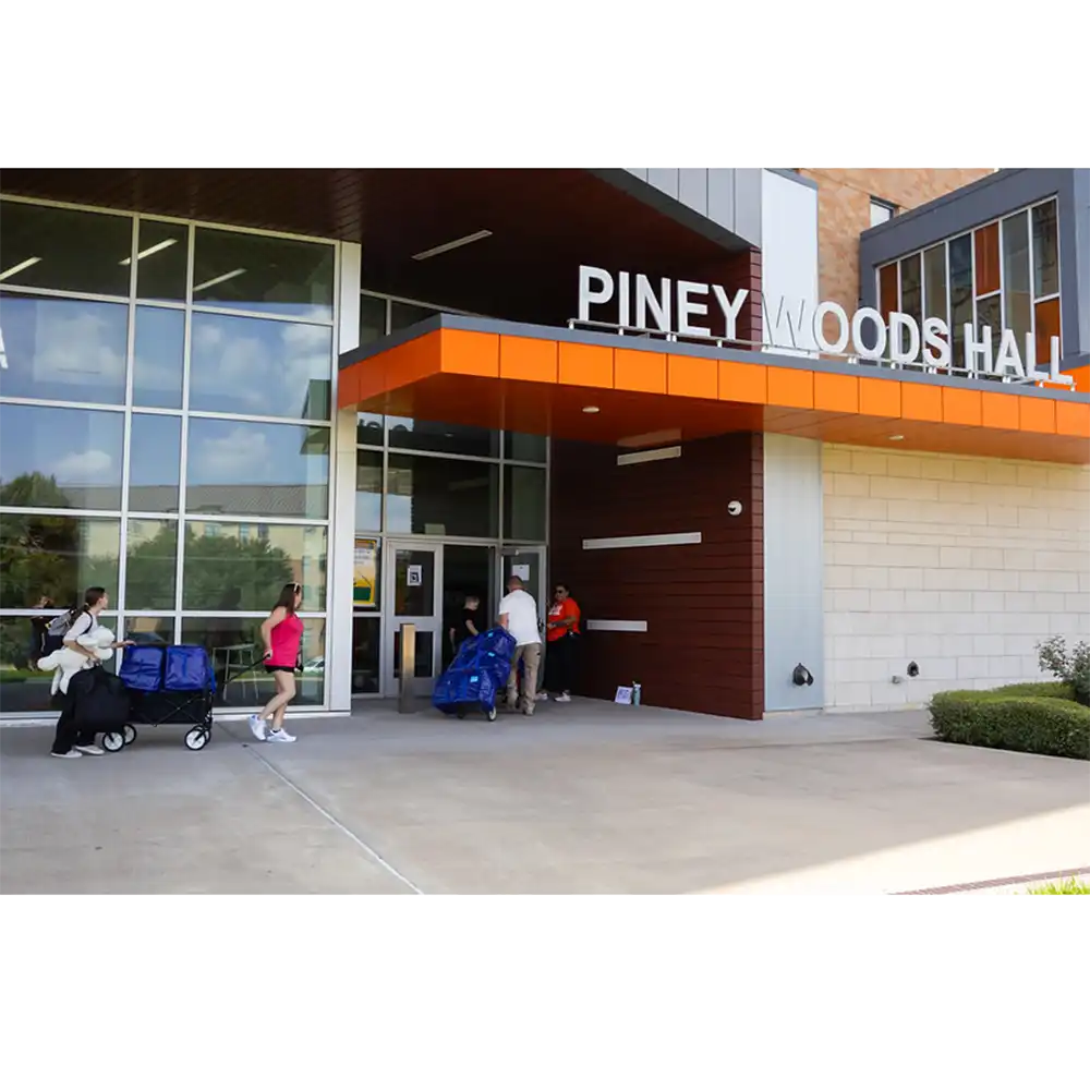 Exterior view of Piney Woods Hall entrance at Sam Houston State University. The building features large glass windows and an orange accent above the doorway with the words “Piney Woods Hall” displayed prominently. Several people are moving items inside using blue rolling carts and carrying bags. The scene suggests a move-in day.