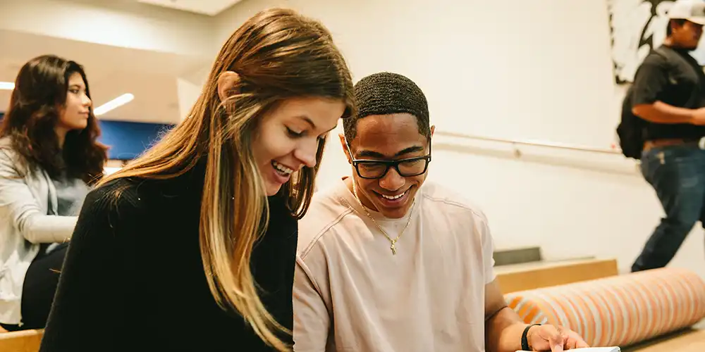 Two smiling SHSU college students sit together indoors, looking at a laptop. Other students are seated and walking in the background.