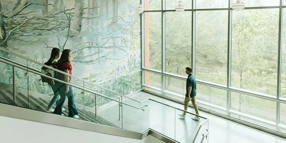 A close-up and wide-angle image of Piney Woods Hall, a modern residence hall located on the campus of Sam Houston State University. The building features a mix of contemporary architectural elements, including large windows, gray metal paneling, and an eye-catching, bright orange overhang at the entrance. The overhang prominently displays the words “PINEY WOODS HALL” in large white block letters. The brick exterior of the building is visible in the background, with rectangular windows evenly spaced across the façade. The photo captures various angles of the building’s entrance, its signage, and parts of the building’s upper floors.
