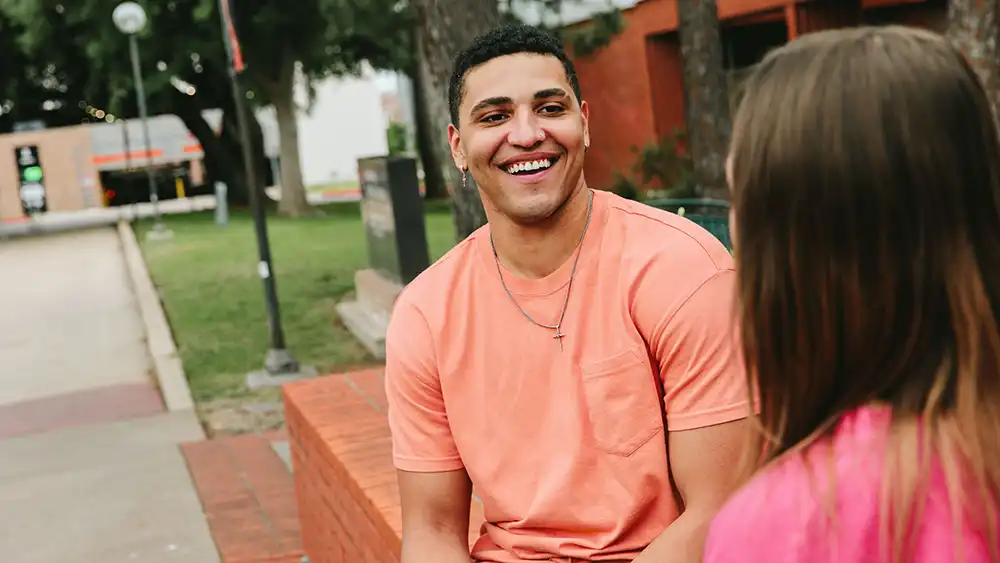 A student in a peach-colored shirt sits on a red brick bench outdoors, smiling and talking to another student in a pink top whose back is to the camera. Trees, a lawn, and apartment buildings are visible in the background, suggesting a college friendly setting.
